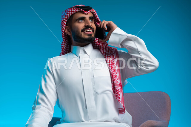 اجراء مكالمة هاتفية عبر الجوال, دردشات ومحادثات عمل مع الزملاء, بورتريه مقرب لشاب عربي سعودي خليجي مبتسم يرتدي الثوب والشماغ التقليدي يجلس على الكرسي يمسك بيده الهاتف المحمول , خلفية زرقاء