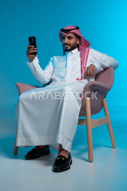 استخدام الهاتف المحمول في التواصل الاجتماعي, دردشة وتواصل عبر الانترنت, بورتريه لشاب عربي سعودي خليجي يرتدي الثوب والشماغ التقليدي يضع يده على الكرسي يمسك بيده الجوال, اجراء مكالمة فيديو مع الاهل والاصدقاء, خلفية زرقاء