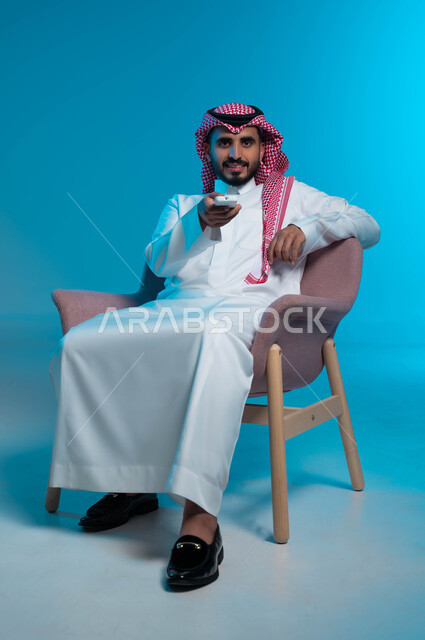 قضاء وقت الفراغ في التسلية والترفيه, مشاهدة الافلام والمسلسلات, بورتريه لشاب عربي سعودي خليجي يرتدي الثوب والشماغ التقليدي يجلس على الكرسي يمسك بيده جهاز التحكم عن بعد بالتلفاز, النظر الى الكاميرا بتعابير المتعة, خلفية زرقاء