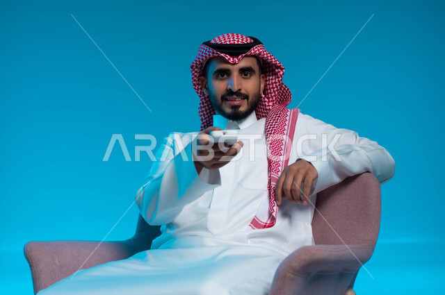 مشاهدة الافلام والمسلسلات, قضاء وقت الفراغ في التسلية والترفيه, بورتريه مقرب لشاب عربي سعودي خليجي يرتدي الثوب والشماغ التقليدي يجلس على الكرسي يمسك بيده جهاز  التحكم عن بعد بالتلفاز, النظر الى الكاميرا بتعابير المتعة, خلفية زرقاء