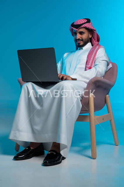 تعابير تدل على الاستمتاع باستخدام الحاسب, دردشة وتواصل عبر الانترنت, استخدام الاجهزة والتقنيات الحديثة والمتطورة, بورتريه لشاب عربي سعودي خليجي مبتسم يرتدي الثوب والشماغ التقليدي يمسك بيده الحاسوب المحمول, خلفية زرقاء