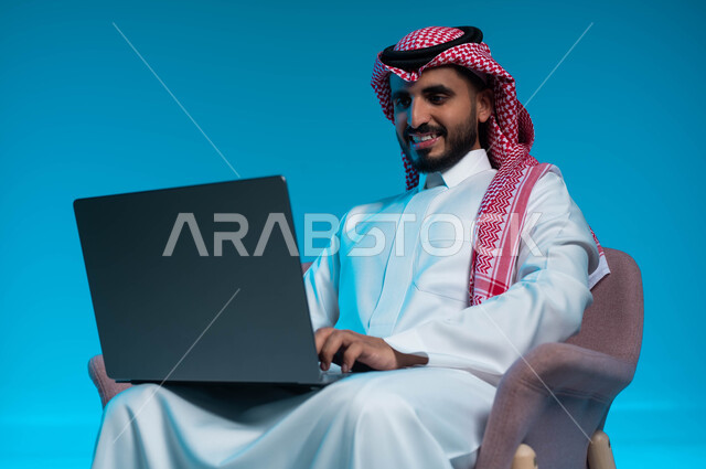 دردشة وتواصل عبر الانترنت, استخدام الاجهزة والتقنيات الحديثة والمتطورة, بورتريه مقرب لشاب عربي سعودي خليجي مبتسم يرتدي الثوب والشماغ التقليدي يمسك بيده الحاسوب المحمول, خلفية زرقاء