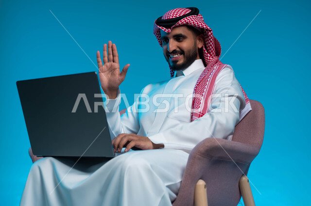 اجراء مكالمة فيديو مع الاهل والاصدقاء عبر الحاسب المحمول, استخدام الاجهزة والتقنيات المتطورة, بورتريه مقرب لشاب عربي سعودي خليجي مبتسم يرتدي الثوب والشماغ التقليدي ويشير بيده على الشاشة, ايماءات التحية والسلام, خلفية زرقاء