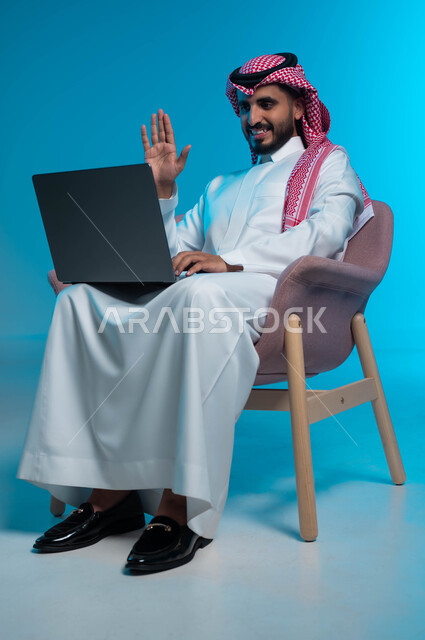 استخدام الاجهزة والتقنيات المتطورة, اجراء مكالمة فيديو عبر الحاسب المحمول, بورتريه لشاب عربي سعودي خليجي يرتدي الثوب والشماغ التقليدي ويشير بيده على الشاشة, ايماءات التحية والسلام, خلفية زرقاء
