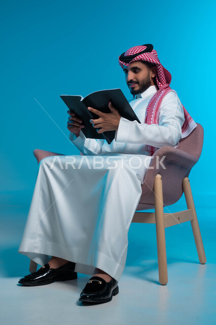 التركيز في احداث القصة المقروءة, مفهوم التعلم الذاتي, ممارسة هواية القراءة والمطالعة, بورتريه لشاب عربي سعودي خليجي يرتدي الثوب والشماغ التقليدي يجلس على الكرسي ويمسك بيده الكتاب, القراءة بتمعن وتفكير, خلفية زرقاء