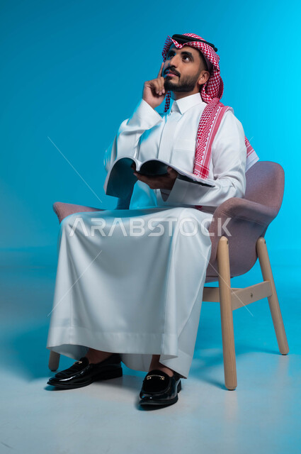 الاستمتاع بمارسة هواية المطالعة, التزود بالعلم والمعرفة, بورتريه لشاب عربي سعودي خليجي يرتدي الثوب والشماغ التقليدي يجلس على الكرسي ويمسك بيده كتاب, النظر الى الاعلى بإيماءات التفكير والتركيز, خلفية زرقاء