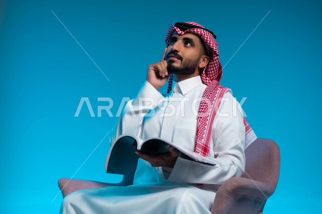 قضاء وقت الفراغ بالتثقيف والتعليم, التزود بالعلم والمعرفة, بورتريه مقرب لشاب عربي سعودي خليجي يرتدي الثوب والشماغ التقليدي يجلس على الكرسي ويحمل بيده كتاب, النظر الى الاعلى بإيماءات التمعن والتركيز, خلفية زرقاء