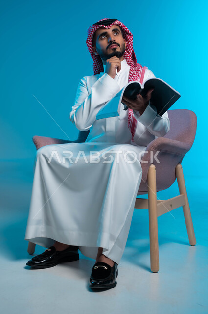Acquiring education and knowledge, contemplating and thinking about the events of the stories read, a portrait of a Saudi Gulf Arab young man wearing the traditional thobe and shemagh, sitting on a chair and holding a book in his hand, looking up carefully and with concentration, blue background