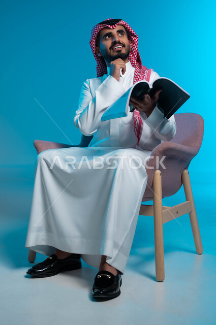 Looking up with gestures of contemplation and concentration, contemplating and thinking about the events of the story being read, practicing the hobby of reading and reading, a close-up portrait of a smiling Saudi Gulf Arab man wearing the traditional thobe and shemagh, sitting on a chair and holding a book in his hand, blue background.