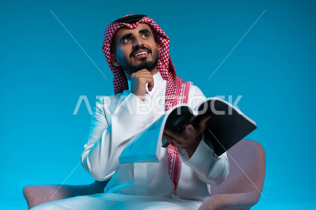 التأمل والتفكير في أحداث القصة المقروءة، ممارسة هواية القراءة والمطالعة، بورتريه مقرب لرجل عربي خليجي سعودي مبتسم يرتدي الثوب والشماغ التقليدي يجلس على الكرسي ويحمل بيده كتاب، النظر الى الأعلى بإيماءات التمعن والتركيز، خلفية زرقاء