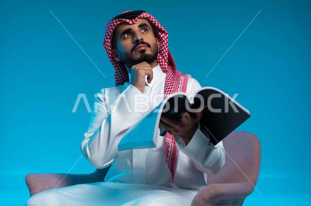 ممارسة هواية القراءة والمطالعة، التأمل والتفكير في أحداث القصة المقروءة، بورتريه لرجل عربي خليجي سعودي يرتدي الثوب والشماغ التقليدي يجلس على الكرسي ويحمل بيده كتاب، النظر الى الأعلى بإيماءات التمعن والتركيز، خلفية زرقاء