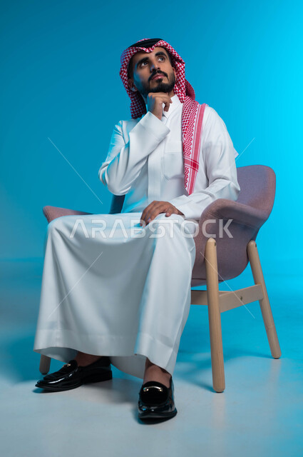 التأمل وشرود الذهن والتخطيط للمستقبل، التفكير لأيجاد حل مشكلة ما، مواجهة المشكلات والتحديات في الحياة العملية، بورتريه لرجل عربي خليجي سعودي يرتدي الثوب والشماغ التقليدي يجلس على الكرسي وينظر الى الأعلى بإيماءات التمعن والتركيز، خلفية زرقاء