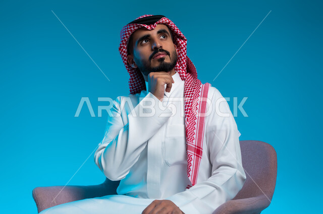 شرود الذهن والتأمل والتخطيط للمستقبل، التفكير لأيجاد حل مشكلة ما، مواجهة المشكلات والتحديات في الحياة العملية، بورتريه لرجل عربي خليجي سعودي يرتدي الثوب والشماغ التقليدي يجلس على الكرسي وينظر الى الأعلى بإيماءات التمعن والتركيز، خلفية زرقاء