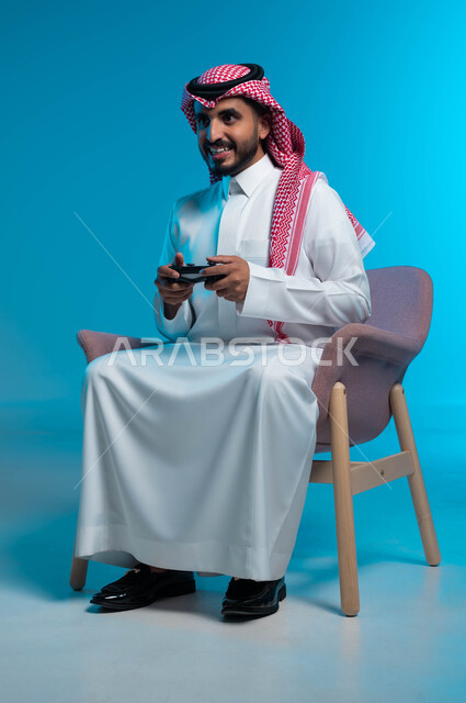 تجربة ترفيهية ممتعة ومثيرة في الألعاب الالكترونية والأجهزة الحديثة، العالم الرقمي الترفيهي، المتعة والتسلية بالفيديو جيمز، بورتريه لشاب عربي خليجي سعودي يرتدي الثوب والشماغ  التقليدي يمسك بيده عصا التحكم بإيماءات التفاعل والاندماج، خلفية زرقاء