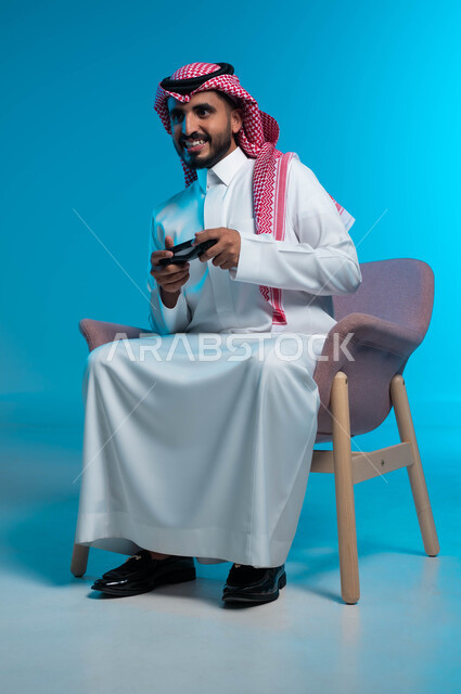 تجربة ترفيهية ممتعة ومثيرة في الألعاب الالكترونية والأجهزة الحديثة، العالم الرقمي الترفيهي، المتعة والتسلية بالفيديو جيمز، بورتريه لشاب عربي خليجي سعودي يرتدي الثوب والشماغ  التقليدي يمسك بيده عصا التحكم بإيماءات التفاعل والاندماج، خلفية زرقاء