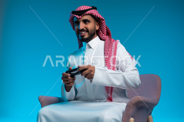 العالم الرقمي الترفيهي، تجربة ترفيهية ممتعة ومثيرة في الألعاب الالكترونية والأجهزة الحديثة، المتعة والتسلية بالفيديو جيمز، بورتريه لشاب عربي خليجي سعودي يرتدي الثوب والشماغ  التقليدي يمسك بيده عصا التحكم بإيماءات التفاعل والاندماج، خلفية زرقاء