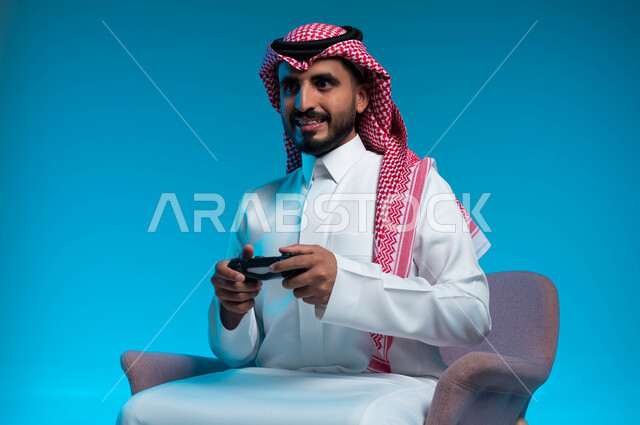 العالم الرقمي الترفيهي، تجربة ترفيهية ممتعة ومثيرة في الألعاب الالكترونية والأجهزة الحديثة، المتعة والتسلية بالفيديو جيمز، بورتريه لشاب عربي خليجي سعودي يرتدي الثوب والشماغ  التقليدي يمسك بيده عصا التحكم بإيماءات التفاعل والاندماج، خلفية زرقاء