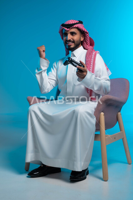 المتعة والتسلية بالفيديو جيمز، التفاعل والاندماج أثناء اللعب بالألعاب الالكترونية الترفيهية، بورتريه لشاب عربي خليجي سعودي مبتسم يرتدي الثوب والشماغ التقليدي يمسك بيده عصا التحكم ويرفع قبضته للأعلى بإيماءات الفوز والانتصار، خلفية زرقاء