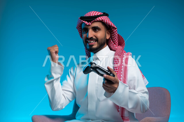 التفاعل والاندماج أثناء اللعب بالألعاب الالكترونية الترفيهية، المتعة والتسلية بالفيديو جيمز، بورتريه مقرب لشاب عربي خليجي سعودي مبتسم يرتدي الثوب والشماغ التقليدي يمسك بيده عصا التحكم ويرفع قبضته للأعلى بإيماءات الفوز والانتصار، خلفية زرقاء