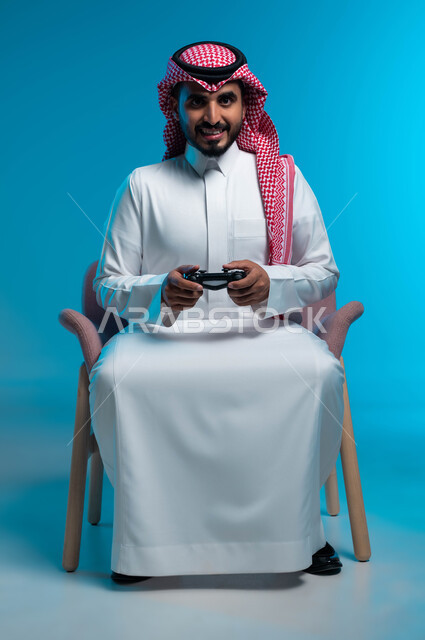 العالم الرقمي الترفيهي، تجربة ترفيهية ممتعة ومثيرة في الألعاب الالكترونية والأجهزة الحديثة، المتعة والتسلية بالفيديو جيمز، بورتريه لشاب عربي خليجي سعودي يرتدي الثوب والشماغ  التقليدي يمسك بيده عصا التحكم بإيماءات التفاعل والاندماج، خلفية زرقاء