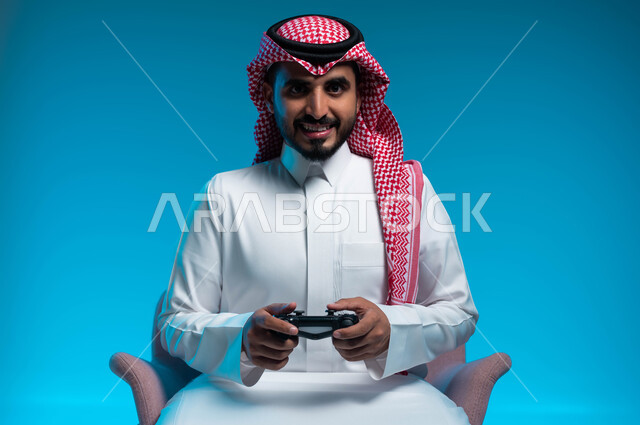 الاستمتاع  باللعب بالألعاب الالكترونية الترفيهية، استخدام الأجهزة التكنولوجية والتقنيات الحديثة، بورتريه لشاب عربي خليجي سعودي مبتسم يرتدي الثوب والشماغ التقليدي يمسك بيده عصا التحكم بإيماءات التفاعل والاندماج، خلفية زرقاء