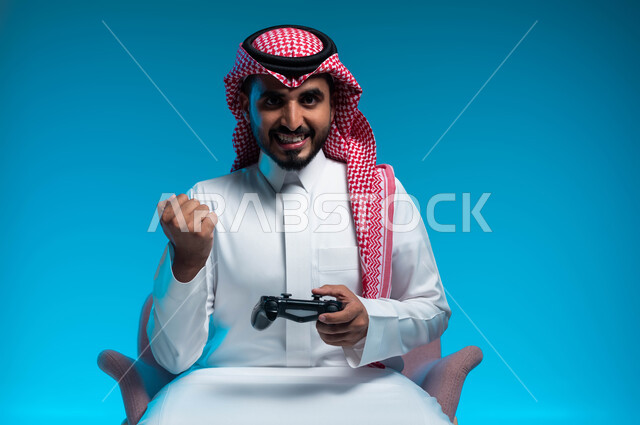 الاندماج والتركيز والتفاعل أثناء اللعب بالألعاب الالكترونية الترفيهية، المتعة والتسلية بالفيديو جيمز، بورتريه مقرب لشاب عربي خليجي سعودي مبتسم يرتدي الثوب والشماغ التقليدي يمسك بيده عصا التحكم ويرفع قبضته للأعلى بإيماءات الفوز والانتصار، خلفية زرقاء
