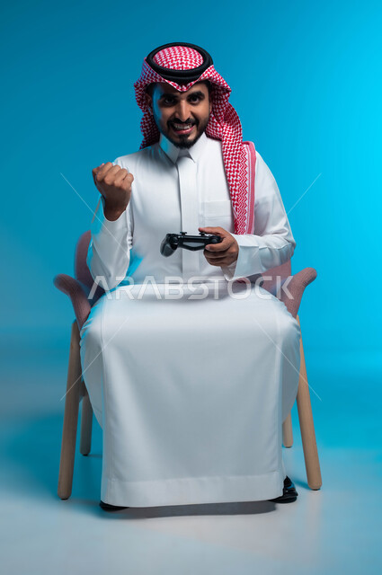 المتعة والتسلية بالفيديو جيمز، الاندماج والتركيز والتفاعل أثناء اللعب بالألعاب الالكترونية الترفيهية، بورتريه لشاب عربي خليجي سعودي مبتسم يرتدي الثوب والشماغ التقليدي يمسك بيده عصا التحكم ويرفع قبضته للأعلى بإيماءات الفوز والانتصار، خلفية زرقاء