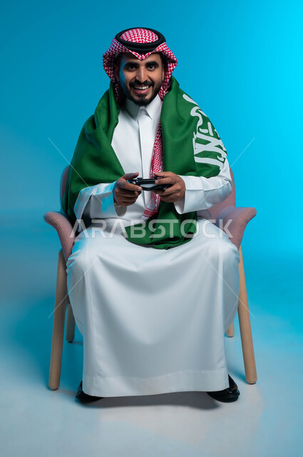 ذكرى اليوم الوطني 23 سبتمبر، الاستمتاع  باللعب بالألعاب الالكترونية الترفيهية، يوم العلم السعودي 11 مارس، بورتريه لشاب عربي خليجي سعودي مبتسم يرتدي الزي التقليدي يضع علم المملمة على كتفيه ويمسك بيده عصا التحكم بإيماءات التفاعل والاندماج، خلفية زرقاء