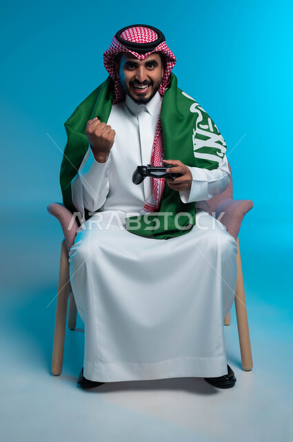 Saudi Flag Day, March 11, commemorating National Day, September 23, enjoying playing entertainment electronic games, portrait of a smiling Saudi Gulf Arab young man wearing traditional clothing, placing the figurine flag on his shoulders, holding a joystick in his hand and raising his fist with gestures of interaction and integration, blue background