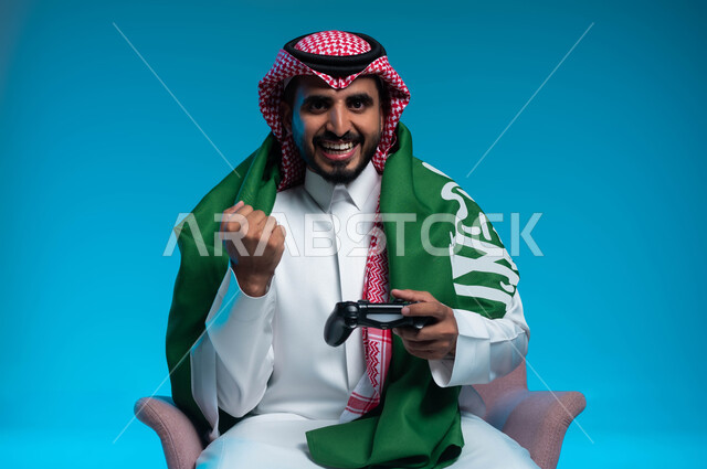 Saudi Flag Day, March 11, commemorating National Day, September 23, enjoying playing entertainment electronic games, portrait of a smiling Saudi Gulf Arab young man wearing traditional clothing, placing the figurine flag on his shoulders, holding a joystick in his hand and raising his fist with gestures of interaction and integration, blue background