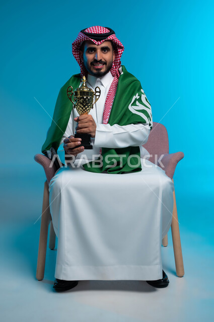 الفخر والاعتزاز بالهوية الوطنية، فوز السعودية بالكأس، اليوم الوطني السعودي 23 سبتمبر، النظر إلى الكاميرا بإيماءات البهجة والفرح بالانتصار، بورتريه لشاب عربي خليجي سعودي مبتسم يرتدي الزي التقليدي وراية الوطن، ذكرى يوم العلم 11 مارس، خلفية زرقاء