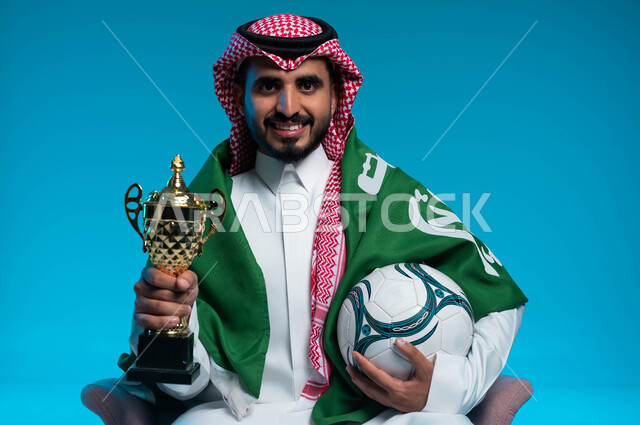 اليوم الوطني السعودي 23 سبتمبر، النظر إلى الكاميرا بإيماءات البهجة والفرح بالانتصارفي مباراة كرة القدم، بورتريه مقرب لشاب عربي خليجي سعودي مبتسم يرتدي الزي التقليدي والراية السعودية يحمل الكرة وكأس الفوز، ذكرى يوم العلم 11 مارس، خلفية زرقاء