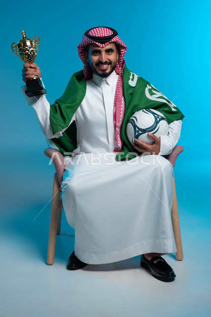 النظر إلى الكاميرا بإيماءات البهجة والفرح بالانتصارفي مباراة كرة القدم، اليوم الوطني السعودي 23 سبتمبر، بورتريه لشاب عربي خليجي سعودي مبتسم يرتدي الزي التقليدي والراية السعودية ويحمل الكرة وكأس الفوز، ذكرى يوم العلم 11 مارس، خلفية زرقاء
