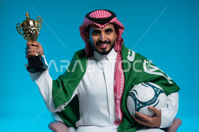اليوم الوطني السعودي 23 سبتمبر، النظر إلى الكاميرا بإيماءات البهجة والفرح بالانتصارفي مباراة كرة القدم، بورتريه مقرب لشاب عربي خليجي سعودي مبتسم يرتدي الزي التقليدي والراية السعودية يحمل الكرة وكأس الفوز، ذكرى يوم العلم 11 مارس، خلفية زرقاء