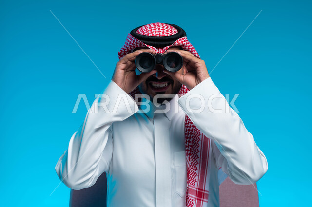 رؤية الاشياء البعيدة عبر التلسكوب، حب الاستكشاف والمغامرة، بورتريه مقرب لرجل عربي خليجي سعودي مبتسم يرتدي الثوب والشماغ التقليدي ينظر من خلال منظار الرؤية ثنائي العينين, التخطيط المستقبلي للأهداف والطموحات، خلفية زرقاء