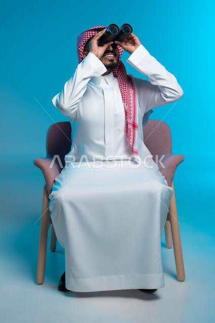 حب الاستكشاف والمغامرة، رؤية الاشياء البعيدة عبر التلسكوب، بورتريه مقرب لرجل عربي خليجي سعودي مبتسم يرتدي الثوب والشماغ التقليدي ينظر من خلال منظار الرؤية ثنائي العينين, التخطيط المستقبلي للأهداف والطموحات، خلفية زرقاء