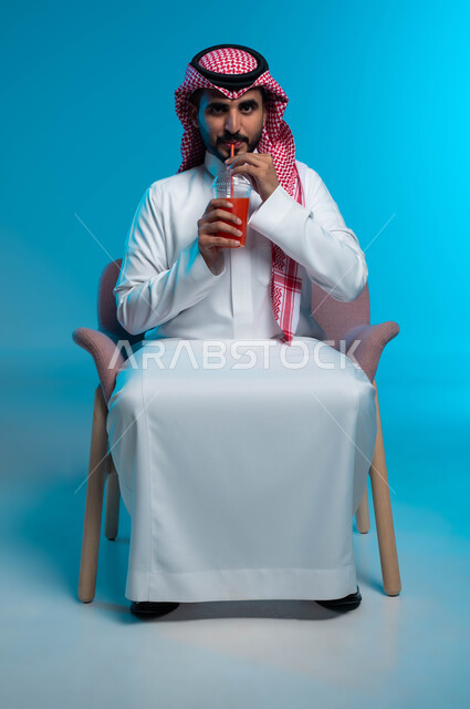 الاستمتاع بتناول المشروبات المفيدة اللذيذة، محاولة التخفيف من حرارة الصيف بالمشروب المنعش، بورتريه لرجل عربي خليجي سعودي مبتسم يرتدي الزي التقليدي يضع سماعة الرأس ويستمتع بشرب العصير الطبيعي البارد، التحديق والنظر إلى الكاميرا، خلفية زرقاء