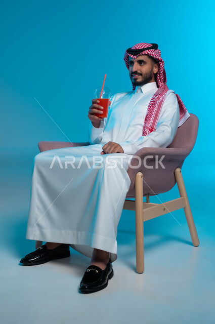 النظر إلى شيء ما بإيماءات السرور، محاولة التخفيف من حرارة الصيف بالعصائر المنعشة، الاستمتاع بتناول المشروبات المفيدة اللذيذة، بورتريه لرجل عربي خليجي سعودي مبتسم يرتدي الزي التقليدي يضع سماعة الرأس ويستمتع بشرب العصير الطبيعي البارد، خلفية زرقاء