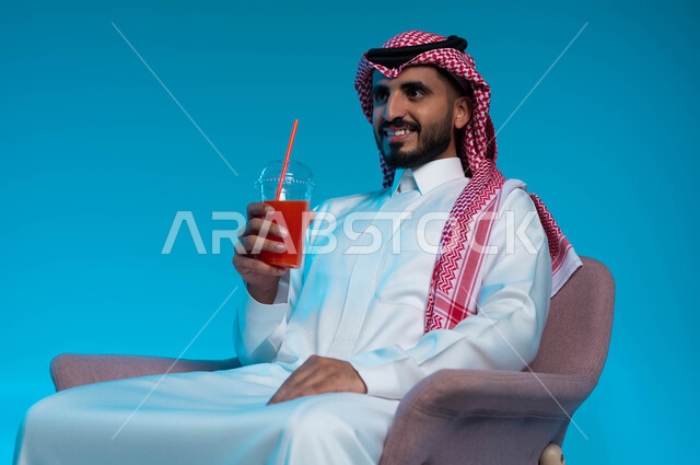 النظر إلى شيء ما بإيماءات السرور، محاولة التخفيف من حرارة الصيف بالعصائر المنعشة، الاستمتاع بتناول المشروبات المفيدة اللذيذة، بورتريه مقرب لرجل عربي خليجي سعودي مبتسم يرتدي الزي التقليدي يضع سماعة الرأس ويستمتع بشرب العصير الطبيعي البارد، خلفية زرقاء