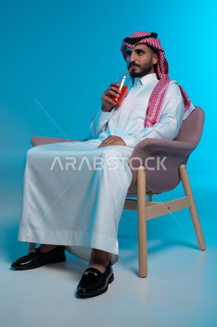 الاستمتاع بتناول المشروبات المفيدة اللذيذة، محاولة التخفيف من حرارة الصيف بالمشروب المنعش، بورتريه لرجل عربي خليجي سعودي مبتسم يرتدي الزي التقليدي يضع سماعة الرأس ويستمتع بشرب العصير الطبيعي البارد، خلفية زرقاء