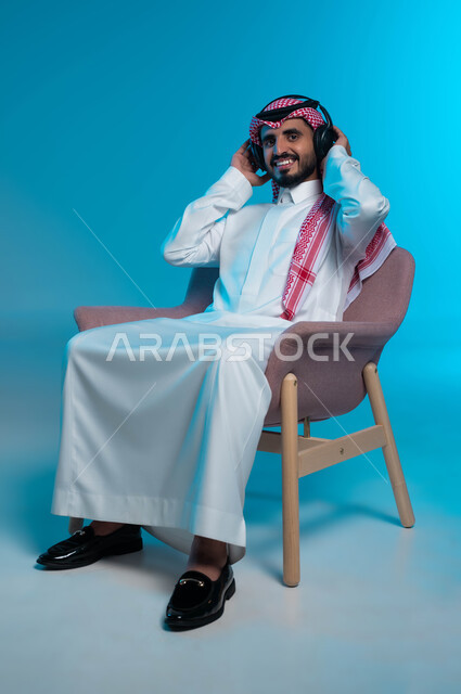 النظر إلى الكاميرا بتعابير السرور، قضاء أوقات ممتعة بالاستماع للمواهب الغنائية، مفهوم الإصغاء والاستمتاع باستخدام الهيدفون، بورتريه لرجل عربي خليجي سعودي مبتسم يرتدي الزي التقليدي يضع سماعة الرأس بإيماءات تدل على الاندماج والطرب، خلفية زرقاء