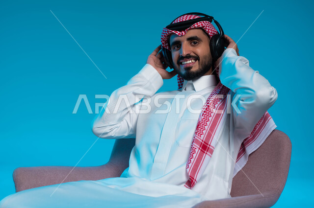 قضاء أوقات ممتعة بالاستماع للمواهب الغنائية، النظر إلى الكاميرا بتعابير السرور، مفهوم الإصغاء والاستمتاع باستخدام الهيدفون، بورتريه مقرب لرجل عربي خليجي سعودي مبتسم يرتدي الزي التقليدي يضع سماعة الرأس بإيماءات الطرب، خلفية زرقاء