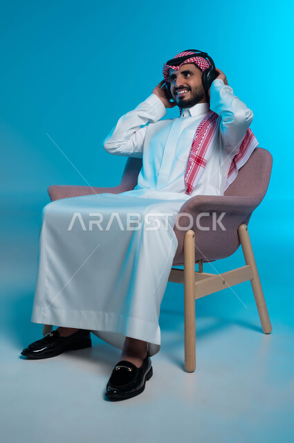 النظر إلى شيء ما بتعابير السرور، قضاء أوقات ممتعة بالاستماع للمواهب الغنائية، مفهوم الإصغاء والاستمتاع باستخدام الهيدفون، بورتريه لرجل عربي خليجي سعودي مبتسم يرتدي الزي التقليدي يضع سماعة الرأس بإيماءات تدل على الاندماج والطرب، خلفية زرقاء