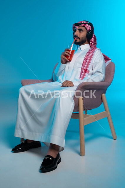 الإصغاء والاستمتاع باستخدام الهيدفون، مشروب صيفي منعش، قضاء أوقات ممتعة بالاستماع للمواهب الغنائية الفنية، بورتريه لرجل عربي خليجي سعودي مبتسم يرتدي الزي التقليدي يضع سماعة الرأس ويستمتع بشرب العصير الطبيعي البارد، خلفية زرقاء