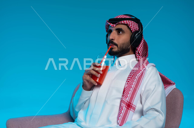 مشروب صيفي منعش، قضاء أوقات ممتعة بالاستماع للمواهب الغنائية الفنية، مفهوم الإصغاء والاستمتاع باستخدام الهيدفون، بورتريه مقرب لرجل عربي خليجي سعودي مبتسم يرتدي الزي التقليدي يضع سماعة الرأس ويستمتع بشرب العصير الطبيعي البارد، خلفية زرقاء
