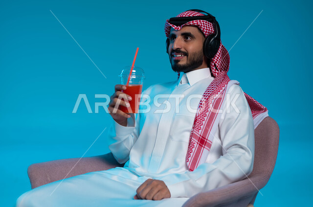 مشروب صيفي منعش، قضاء أوقات ممتعة بالاستماع للمواهب الغنائية الفنية، مفهوم الإصغاء والاستمتاع باستخدام الهيدفون، بورتريه مقرب لرجل عربي خليجي سعودي مبتسم يرتدي الزي التقليدي يضع سماعة الرأس ويستمتع بشرب العصير الطبيعي البارد، خلفية زرقاء