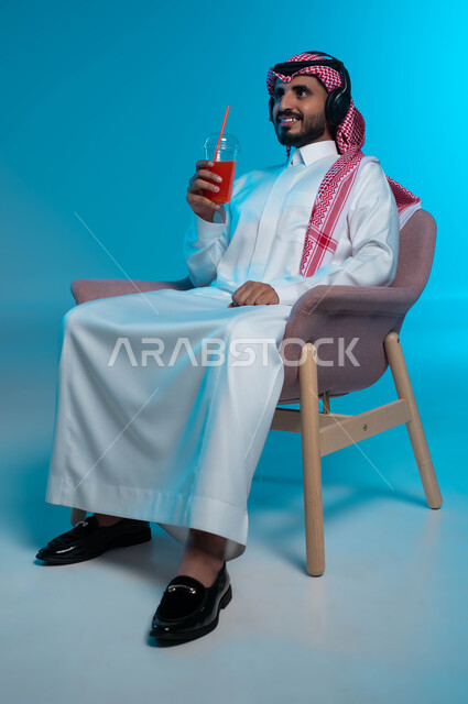 الإصغاء والاستمتاع باستخدام الهيدفون، مشروب صيفي منعش، قضاء أوقات ممتعة بالاستماع للمواهب الغنائية الفنية، بورتريه لرجل عربي خليجي سعودي مبتسم يرتدي الزي التقليدي يضع سماعة الرأس ويستمتع بشرب العصير الطبيعي البارد، خلفية زرقاء