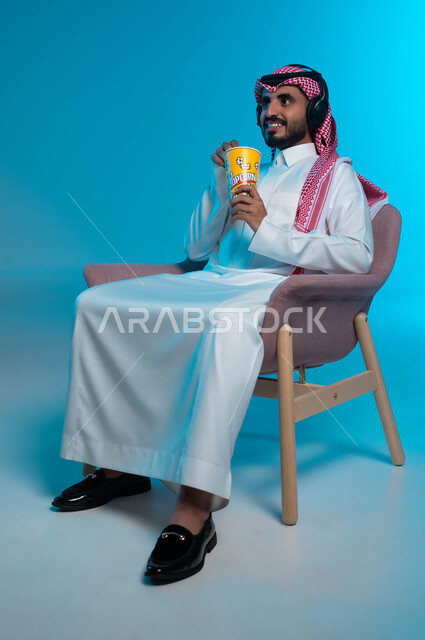 قضاء أوقات ممتعة ومسلية، الإصغاء والاستمتاع باستخدام الهيدفون، الاستماع إلى المواهب الغنائية الفنية، بورتريه لرجل عربي خليجي سعودي مبتسم يرتدي الزي التقليدي يضع سماعة الرأس ويستمتع بتناول الفوشار، خلفية زرقاء