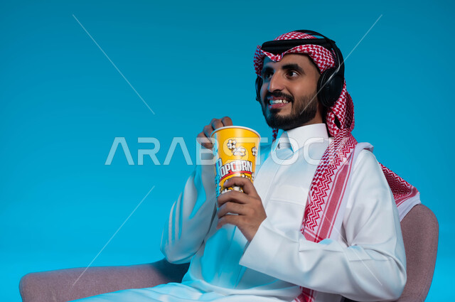 Listening to songs and audio clips, spending fun and entertaining times, listening and enjoying using headphones, listening to artistic singing talents, a close-up portrait of a smiling Saudi Gulf Arab man wearing traditional clothing, wearing headphones and enjoying eating popcorn, blue background.