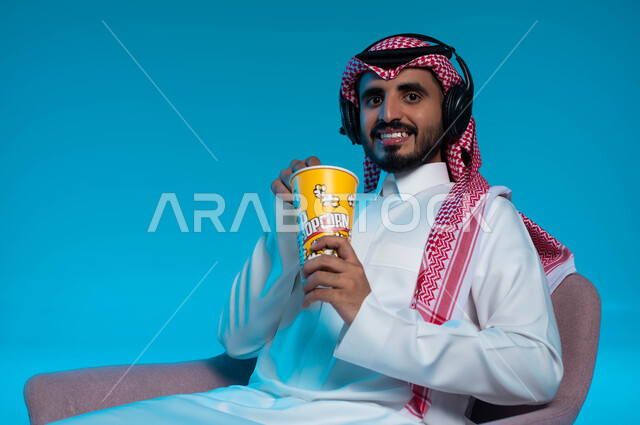 Listening to songs and audio clips, spending fun and entertaining times, listening and enjoying using headphones, listening to artistic singing talents, a close-up portrait of a smiling Saudi Gulf Arab man wearing traditional clothing, wearing headphones and enjoying eating popcorn, blue background.